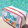 White pouch with 'Hello Weekend' text in colorful letters, placed on a sandy surface with a pool in the background.