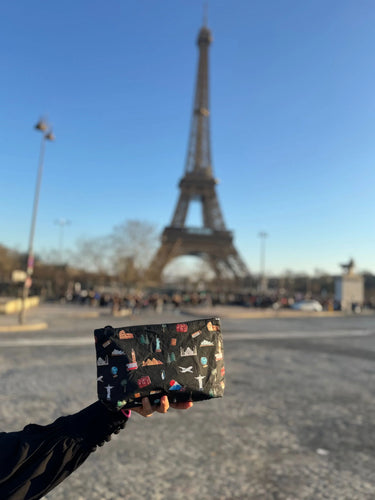 A cosmetic zipper pouch in black with world landmark pictures printed onto it being held in front of the Eiffel Tower 