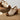 A pair of women's open-toe block-heel sandals with metallic champagne-gold textured uppers, bronze-tone metal studs, and brown wooden soles, displayed on a rustic wooden surface with a light gray background.