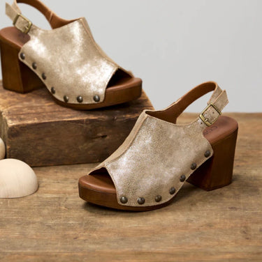 A pair of women's open-toe block-heel sandals with metallic champagne-gold textured uppers, bronze-tone metal studs, and brown wooden soles, displayed on a rustic wooden surface with a light gray background.