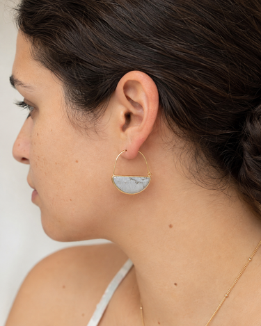 A close-up side profile of a woman wearing a gold hoop earring with a semi-circular light gray and white marbled stone insert.