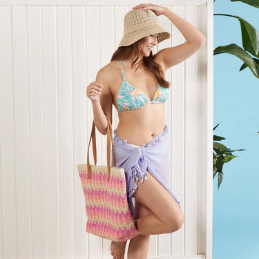 Woman in a tropical-themed bikini with a straw hat and colorful bag against a white wooden background.