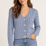 A woman wearing an acid wash cable knit cardigan in denim blue with blue jeans on a white background