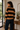 A woman wearing a tan and black horizontally striped knit sweater and dark blue jeans, standing in a warm, rustic indoor setting with a window, potted plants, and a wooden console table.