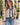 A woman wearing a beige quilted jacket with a blue denim collar, cuffs, and front placket, styled with a white top and blue jeans, standing outdoors on a paved walkway.