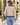 A woman viewed from behind wearing a quilted beige jacket with contrasting blue denim trim on the collar, cuffs, and hem, paired with light blue jeans, set against a blurred outdoor storefront background.