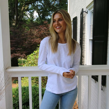 Woman in a white long-sleeve shirt and light blue jeans standing on a porch.