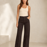 A woman modeling a pair of dark brown wide-leg jeans with a cream tank top, standing in a minimalist beige studio setting.