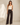 A woman modeling a pair of dark brown wide-leg jeans with a cream tank top, standing in a minimalist beige studio setting.