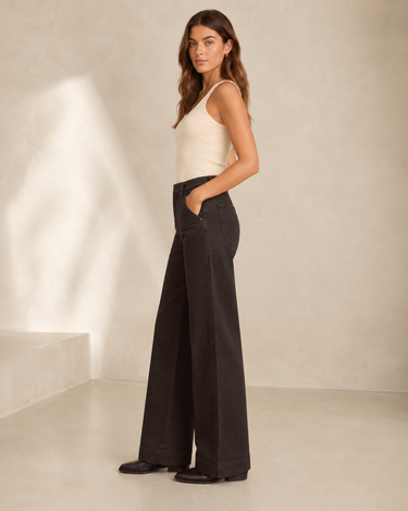 A woman stands in profile wearing a cream ribbed tank top and high-waisted, wide-leg black pants against a warm beige textured studio wall.
