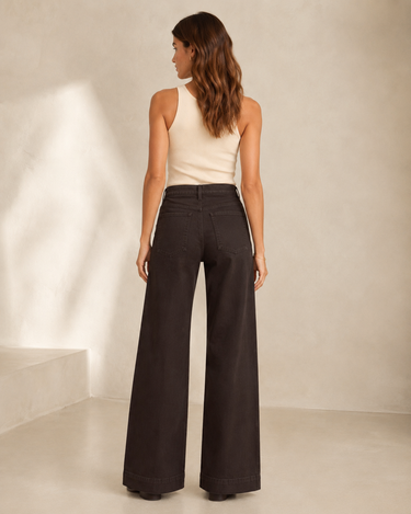 A woman shown from the back wearing dark brown wide-leg pants and a cream sleeveless top, standing in a minimalist beige studio setting.