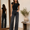 A woman modeling a black short-sleeve crewneck t-shirt and dark gray wide-leg jeans, standing in a brightly lit room with a large mirror showing her back, a wooden bench, and a potted plant.
