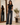 A woman modeling a black short-sleeve crewneck t-shirt and dark gray wide-leg jeans, standing in a brightly lit room with a large mirror showing her back, a wooden bench, and a potted plant.