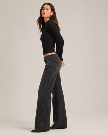 A woman modeling a black long-sleeve crop top and dark charcoal gray wide-leg jeans against a plain light gray studio background.