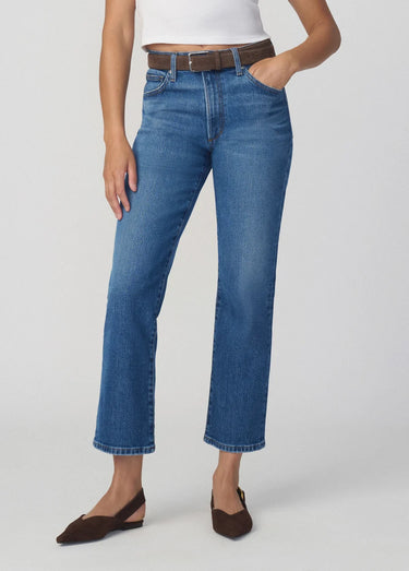 A model wearing a pair of medium-wash blue cropped jeans with a brown belt, paired with a white top and brown slingback flats, against a light gray studio background.