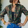 A woman wearing a vintage restructured men’s blouse in camo with white jeans on a neutral background.