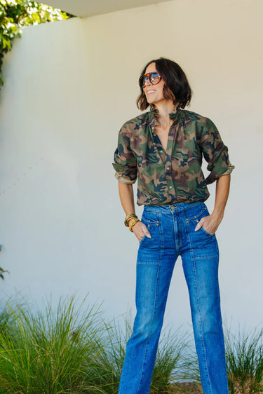 A woman wearing a vintage restructured men’s blouse in camo with white jeans on a neutral background.