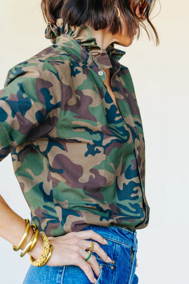 A woman wearing a vintage restructured men’s blouse in camo with white jeans on a neutral background.