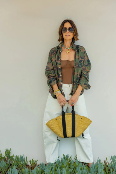 A woman wearing a vintage restructured men’s blouse in camo with white jeans on a neutral background.