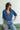 Woman wearing a blue denim shirt and jeans with sunglasses, standing against a light background.