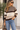 A woman wearing a brown, beige, and cream striped knit sweater with blue jeans, sunglasses, and holding a brown handbag, standing in front of a modern building.