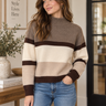 A woman wearing a cozy knit sweater with horizontal stripes in taupe, cream, and dark brown, paired with blue jeans in a warm, modern indoor setting.