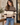 A woman wearing a cozy knit sweater with horizontal stripes in taupe, cream, and dark brown, paired with blue jeans in a warm, modern indoor setting.
