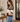 A woman wearing a color-block knit sweater with beige, cream, and dark brown horizontal stripes, paired with blue jeans, standing in a cozy, modern room.