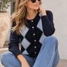 A woman wearing a navy and light blue argyle cardigan sweater, blue jeans, and sunglasses sits on stone steps in front of a white stucco wall.