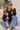 A woman wearing a navy and light blue argyle cardigan sweater, blue jeans, and sunglasses sits on stone steps in front of a white stucco wall.