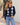 A woman wearing a navy blue cardigan with a light blue and white argyle pattern, paired with light blue jeans, standing outdoors against a white stucco wall.