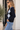 A woman wearing a navy blue and light gray argyle cardigan with light blue jeans, standing in front of a light-colored wall next to a potted plant.