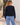 A woman viewed from behind wearing a dark navy knit sweater and light blue jeans, standing outdoors against a light-colored stucco wall.