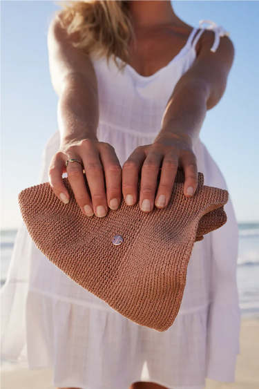 Person holding a brown woven clutch bag on a beach