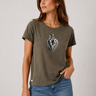 A woman wearing a short-sleeve crewneck t-shirt in olive green with a black and white graphic of a heart and cross on the chest, paired with blue jeans, photographed against a neutral light gray background.