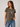 A woman wearing a short-sleeve crewneck t-shirt in olive green with a black and white graphic of a heart and cross on the chest, paired with blue jeans, photographed against a neutral light gray background.