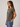 A woman wearing an olive green short-sleeve t-shirt with a black and white graphic print on the left chest, paired with blue jeans, photographed against a neutral light gray background.