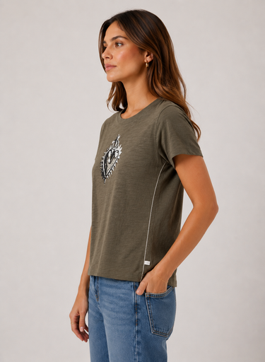 A woman wearing an olive green short-sleeve t-shirt with a black and white graphic print on the left chest, paired with blue jeans, photographed against a neutral light gray background.