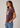 A woman wearing a short-sleeve crewneck t-shirt in a washed dark plum purple with a white graphic print on the front, paired with blue jeans, photographed against a neutral light gray background.