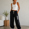 A woman modeling black jogger sweatpants with three white heart graphics on the leg, paired with a white ribbed tank top and white sneakers in a bright, modern room.