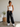 A woman modeling black jogger sweatpants with three white heart graphics on the leg, paired with a white ribbed tank top and white sneakers in a bright, modern room.
