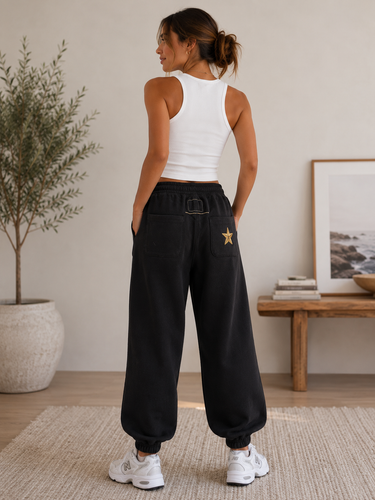 A woman stands with her back to the camera wearing black wide-leg sweatpants with a small gold star patch on the pocket, a white ribbed tank top, and white sneakers in a bright indoor room.