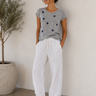 A woman stands against a white wall wearing a heather gray short-sleeve t-shirt with scattered navy blue star motifs, paired with loose white drawstring pants and white sneakers. A potted olive tree is visible on the left.