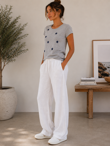 A woman stands in a bright indoor room wearing a light gray short-sleeve t-shirt with small dark star motifs and loose white wide-leg pants.