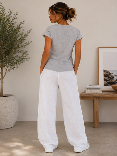 A woman shown from the back wearing a heather gray short-sleeve t-shirt and white wide-leg pants, standing in a minimalist room with a potted plant and a wooden side table.
