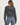 A woman shown from the back wearing a dark gray cable-knit cropped hoodie with a hood, ribbed cuffs and hem, a small red logo patch on the hood, and a gold star patch on the sleeve, paired with blue jeans against a light gray studio background.