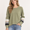 A woman wearing an olive green crewneck sweatshirt with white star details on the sleeves and striped cuffs, paired with blue jeans, against a plain light background.