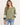 A woman wearing an olive green crewneck sweatshirt with white star details on the sleeves and striped cuffs, paired with blue jeans, against a plain light background.