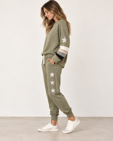 A woman modeling an olive green lounge set consisting of a crewneck sweatshirt and matching joggers, both decorated with white star patches. The sweatshirt features striped details on the sleeves in cream, gray, and black.