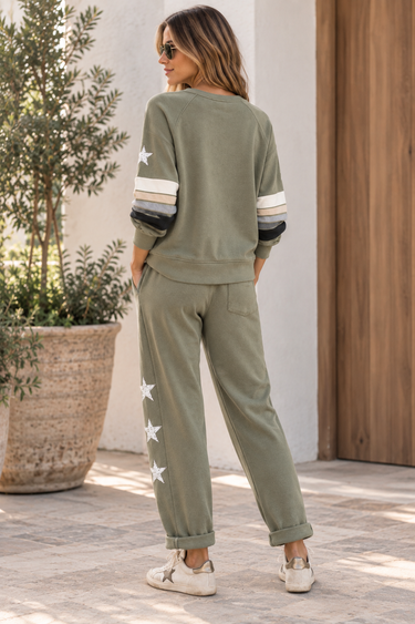 A woman stands outdoors wearing a matching sage green lounge set with white star details on the sleeves and legs, paired with white sneakers.